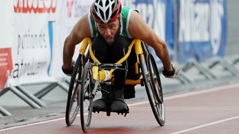 Mohamed Berrahal A man in a yellow racing chair pushes forward in the outside lane of a race.