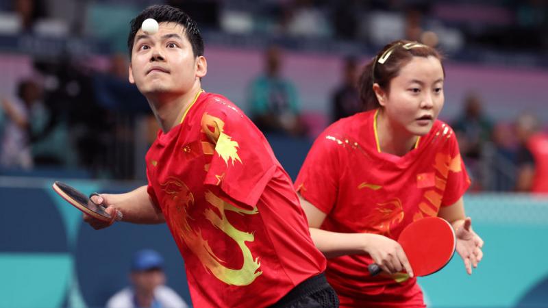 Two athletes play table tennis on the same team, a man looks at the ball and prepares to hit it, a woman braces herself to prepare for the next play